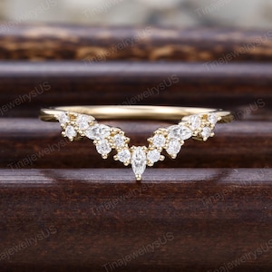 May include: A gold V-shaped ring featuring a series of small, clear, round-cut stones and a single marquise-cut stone at the center point. The band is thin and polished, with the stones arranged in a curved, symmetrical pattern along the top.