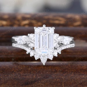 May include: A white gold engagement ring with an emerald-cut center stone, flanked by smaller pear-shaped and round diamonds. The band features additional pav&eacute;-set diamonds, creating a sparkling, elegant design.