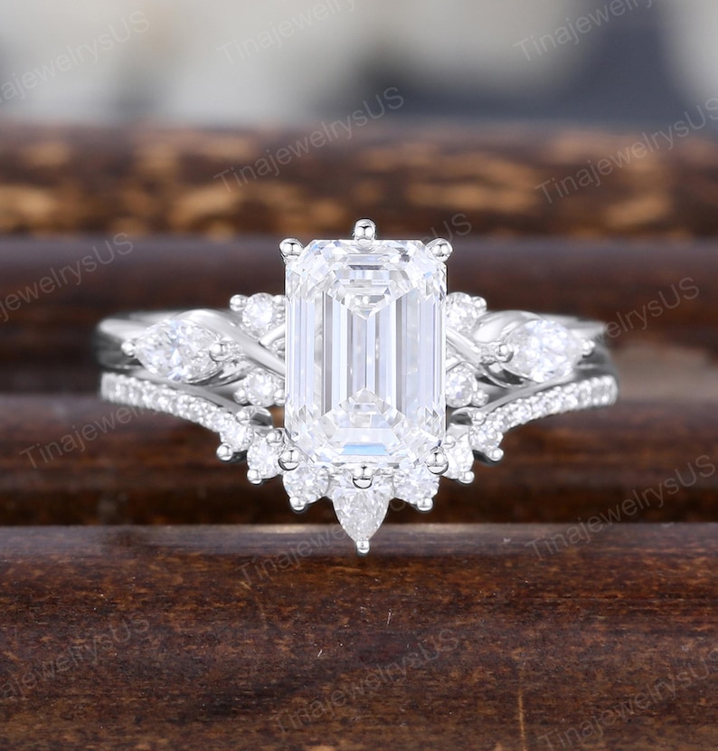 May include: A white gold engagement ring with an emerald-cut center stone, flanked by smaller pear-shaped and round diamonds. The band features additional pav&eacute;-set diamonds, creating a sparkling, elegant design.