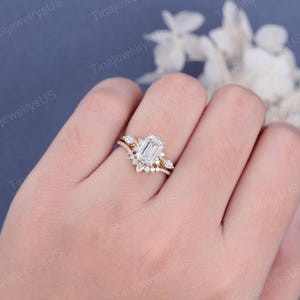 May include: A gold engagement ring set featuring an emerald-cut diamond center stone and a curved band with smaller accent stones. The ring is set on a hand, showcasing the intricate design and sparkle.