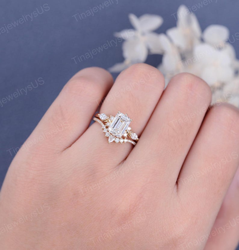 May include: A gold engagement ring set featuring an emerald-cut diamond center stone and a curved band with smaller accent stones. The ring is set on a hand, showcasing the intricate design and sparkle.