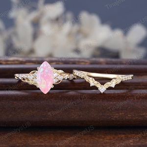 以下が含まれることがあります： マーキス型のピンクオパールをあしらったゴールドトーンのリングセット。婚約指輪は葉とダイヤモンドのデザインで、結婚指輪は葉とダイヤモンドのアクセントが付いた曲線的なバンドです。リングは濃い木製の表面に展示されています。