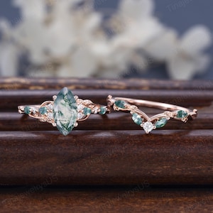 May include: A rose gold engagement ring and wedding band set. The engagement ring features a marquise-cut moss agate center stone, surrounded by small, round, and marquise-cut gemstones. The wedding band has a curved design with matching gemstones.