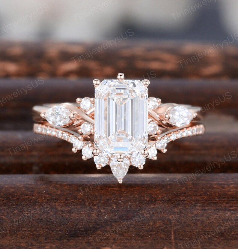 May include: A rose gold engagement ring set featuring an emerald-cut center stone and a delicate band. The set includes a matching band with pear-shaped and round accent stones, creating a sparkling and elegant design.