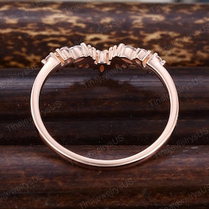 Puede incluir: Anillo de oro rosa con un dise&ntilde;o curvo. El anillo presenta una fila de peque&ntilde;as piedras preciosas transparentes a lo largo de la curva superior. La banda es lisa y pulida. El anillo est&aacute; sobre un fondo oscuro y texturizado.