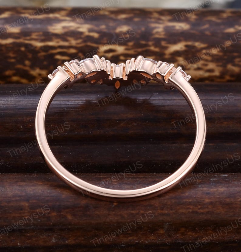 Puede incluir: Anillo de oro rosa con un dise&ntilde;o curvo. El anillo presenta una fila de peque&ntilde;as piedras preciosas transparentes a lo largo de la curva superior. La banda es lisa y pulida. El anillo est&aacute; sobre un fondo oscuro y texturizado.