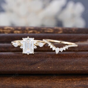 May include: A gold engagement ring set featuring an emerald-cut center stone and a curved band. The main ring has side stones and a delicate design. The set is displayed on a dark wooden surface.