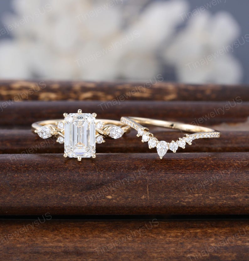 May include: A gold engagement ring set featuring an emerald-cut center stone and a curved band. The main ring has side stones and a delicate design. The set is displayed on a dark wooden surface.
