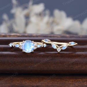 Puede incluir: Un juego de anillos dorados con una piedra preciosa azul claro grande y redonda y piedras claras más pequeñas. El juego incluye una banda curva con piedras preciosas azules a juego. Los anillos se exhiben sobre una superficie de madera oscura.