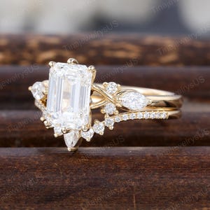 May include: A gold engagement ring set with a large, rectangular, clear gemstone as the centerpiece. The ring features smaller, clear accent stones and a delicate band. The ring is set against a dark, wooden background.