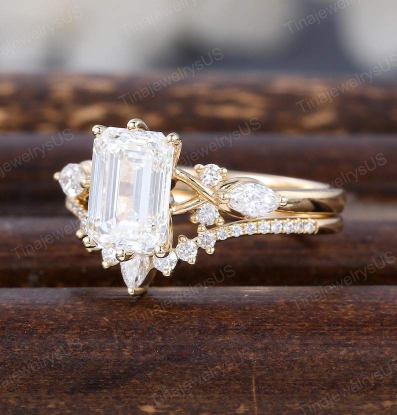 May include: A gold engagement ring set with a large, rectangular, clear gemstone as the centerpiece. The ring features smaller, clear accent stones and a delicate band. The ring is set against a dark, wooden background.