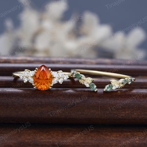 Może przedstawiać: Złoty zestaw pierścionków z pomarańczowym kamieniem szlachetnym w kształcie łzy, otoczonym białymi kamieniami akcentującymi. Zestaw zawiera pasującą obrączkę z zielonymi i białymi kamieniami szlachetnymi oraz detalami w kształcie liści. Pierścionki są prezentowane na ciemnej drewnianej powierzchni.
