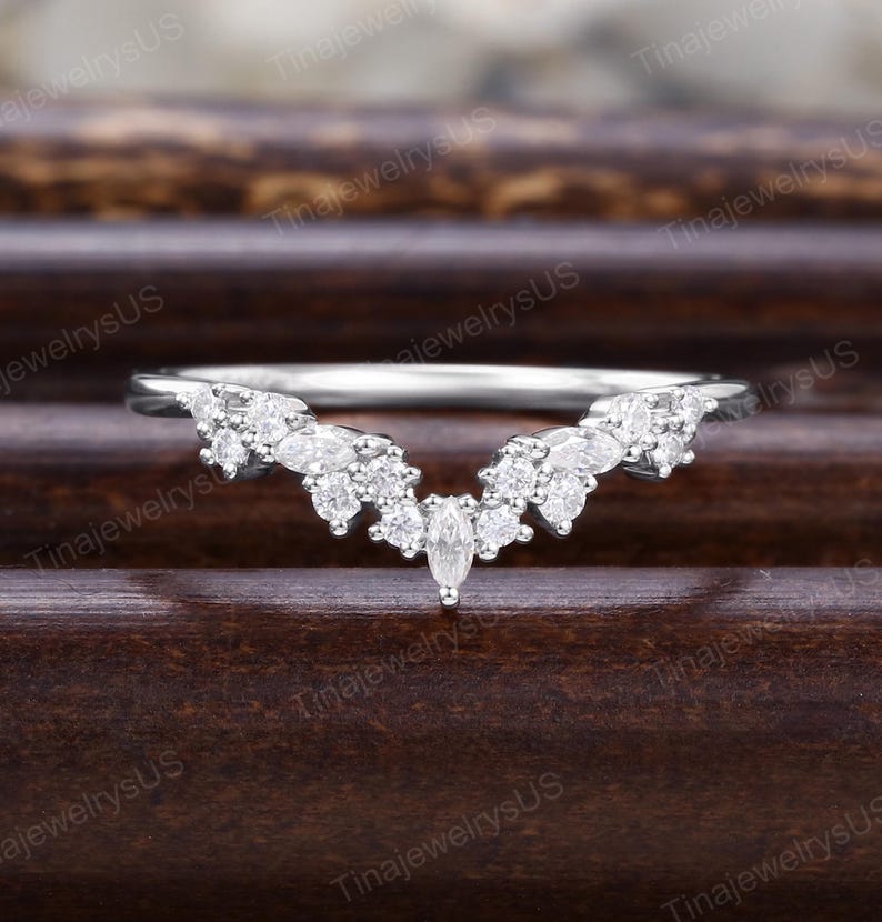 Puede incluir: Un anillo plateado con un dise&ntilde;o curvo, adornado con m&uacute;ltiples piedras preciosas claras y brillantes. El anillo presenta una piedra central con corte marquesa, rodeada de piedras m&aacute;s peque&ntilde;as, de corte redondo, creando una apariencia delicada y elegante.