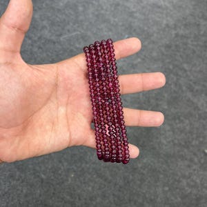 May include: A strand of deep red gemstone beads. The beads are round and smooth, and they are strung on a clear thread. The beads are arranged in a straight line, and they are all the same size.