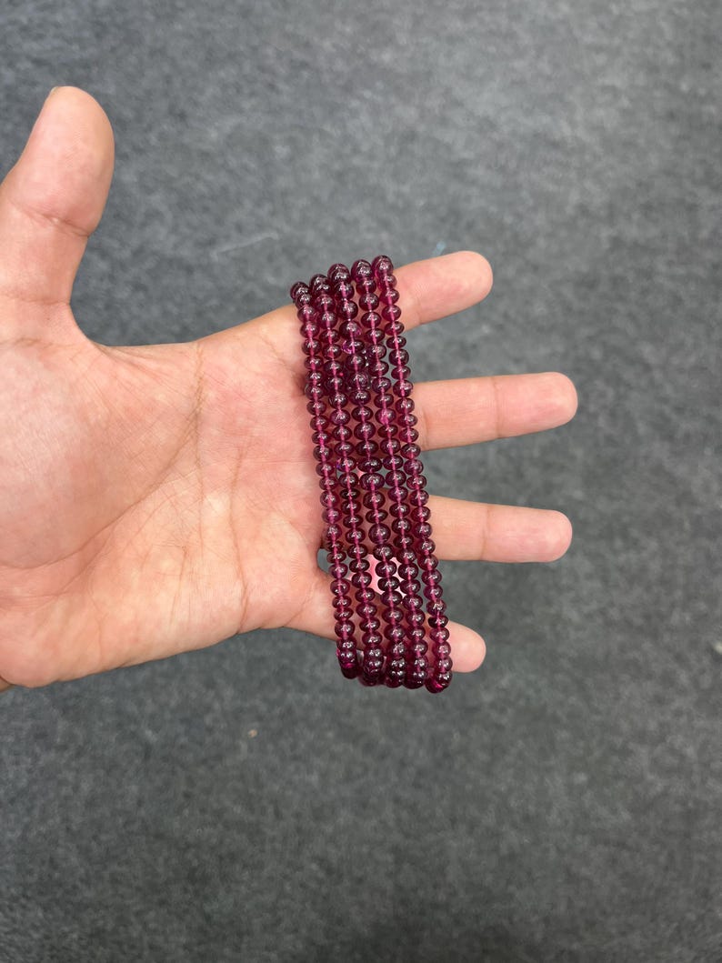 May include: A strand of deep red gemstone beads. The beads are round and smooth, and they are strung on a clear thread. The beads are arranged in a straight line, and they are all the same size.