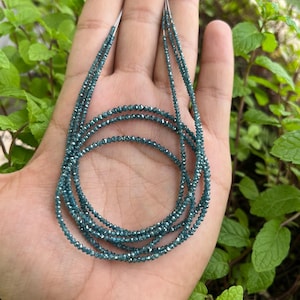 K&ouml;nnte beinhalten: Eine Kette aus blauen Diamantperlen, auf einem silbernen Faden aufgef&auml;delt, wird auf einer Hand gezeigt. Die Perlen sind klein und rund und haben ein funkelndes, facettiertes Aussehen.