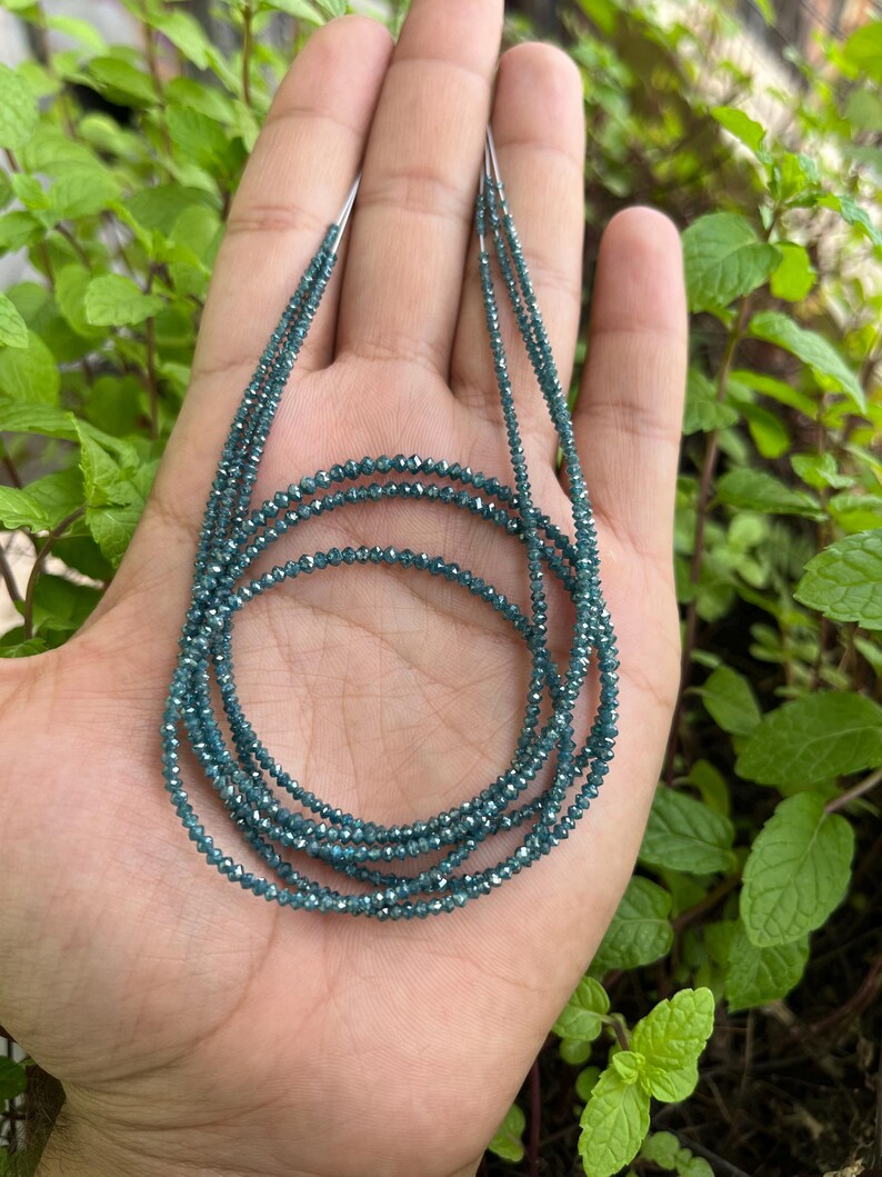 K&ouml;nnte beinhalten: Eine Kette aus blauen Diamantperlen, auf einem silbernen Faden aufgef&auml;delt, wird auf einer Hand gezeigt. Die Perlen sind klein und rund und haben ein funkelndes, facettiertes Aussehen.