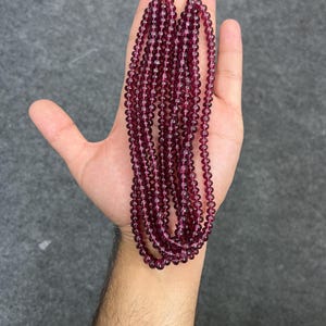 May include: A strand of deep purple gemstone beads, likely amethyst or garnet, strung on a clear thread. The beads are round and smooth, and the strand is long enough to be worn as a necklace or bracelet.