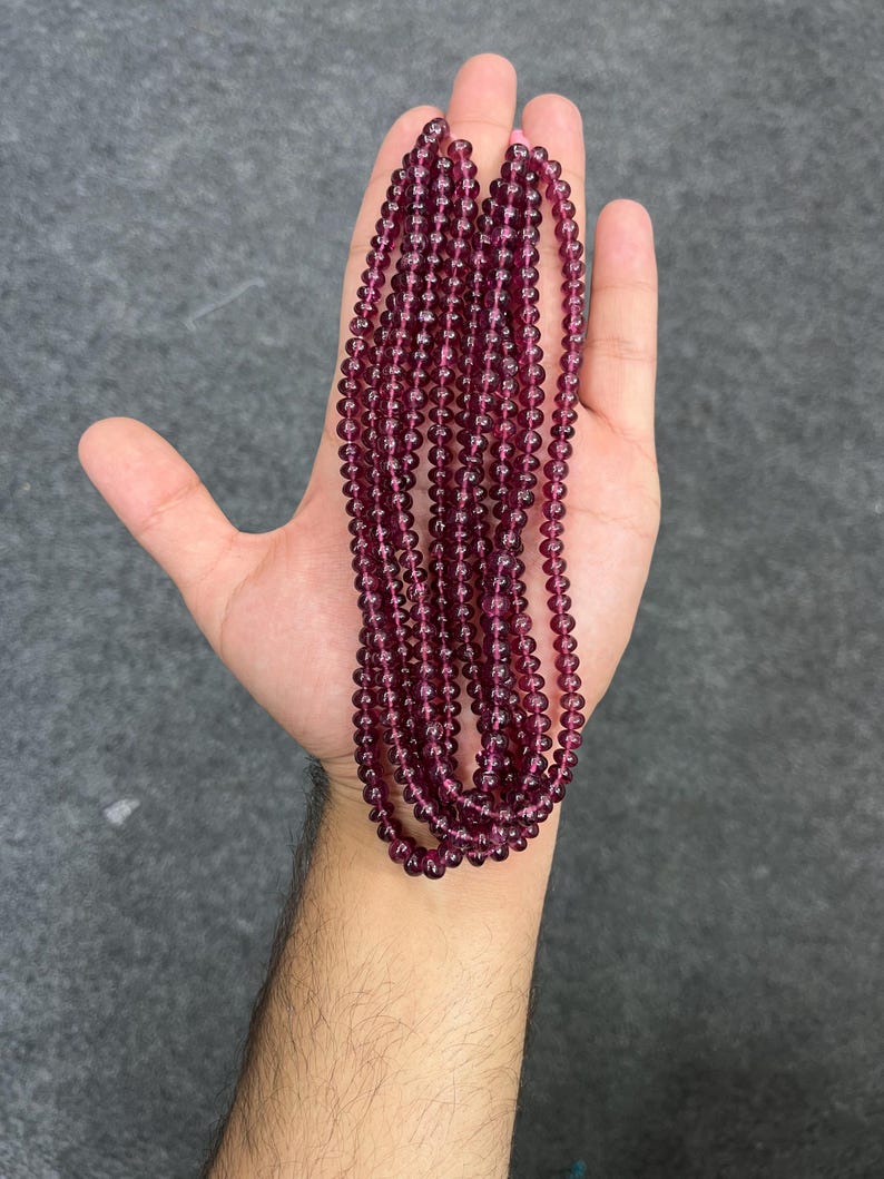 May include: A strand of deep purple gemstone beads, likely amethyst or garnet, strung on a clear thread. The beads are round and smooth, and the strand is long enough to be worn as a necklace or bracelet.