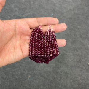May include: A strand of deep red gemstone beads, likely garnet, strung on a thin pink thread. The beads are round and uniform in size, creating a smooth and elegant look.