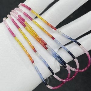 Puede incluir: Un collar de múltiples hilos con pequeñas cuentas de piedras preciosas facetadas en un degradado de colores vibrantes. Las cuentas transicionan a través de tonos rosa, amarillo, naranja, rojo, azul claro y azul oscuro, creando un espectro continuo.