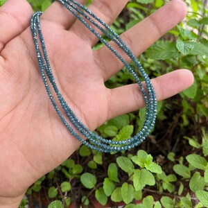 K&ouml;nnte beinhalten: Eine Halskette aus mehreren Reihen kleiner, dunkelblauer, facettierter Perlen. Die Halskette wird auf einer Hand vor einem Hintergrund aus gr&uuml;nem Laub gezeigt.
