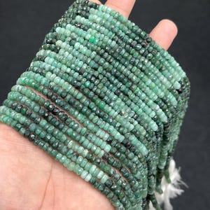 Peut inclure: Plusieurs brins de perles d'émeraude facettées de différentes nuances de vert. Les perles sont petites et uniformes, enfilées ensemble pour créer un aspect fluide et texturé. Les perles sont tenues dans une main, avec des pompons blancs à l'extrémité des brins.