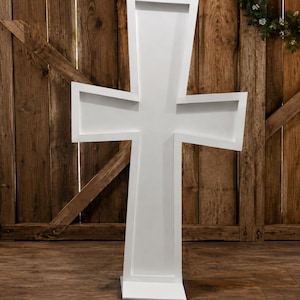 May include: A large, freestanding white cross with a recessed center panel and a flat base. The structure features clean, straight lines and a smooth, matte finish, designed to stand upright on a level surface as a decorative display piece.