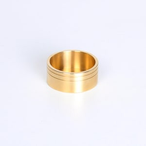 Op de afbeelding: Een goudkleurige ring met een cilindrische vorm. De ring heeft twee dunne, horizontale groeven aan de buitenkant. De binnenkant van de ring is hol. De ring staat tegen een witte achtergrond.