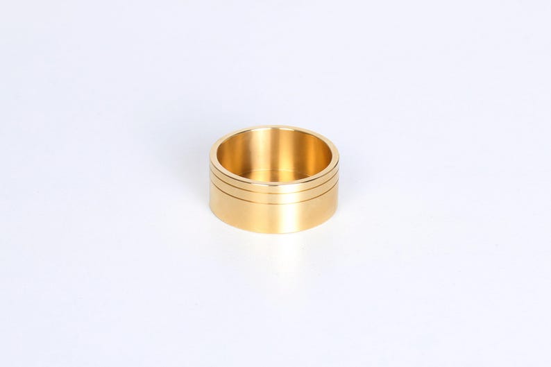 Op de afbeelding: Een goudkleurige ring met een cilindrische vorm. De ring heeft twee dunne, horizontale groeven aan de buitenkant. De binnenkant van de ring is hol. De ring staat tegen een witte achtergrond.