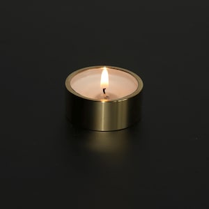 Op de afbeelding: Een brandende waxinelichtkaars in een goudkleurige metalen houder. De kaars heeft een witte was binnenkant en een kleine, flikkerende vlam. De houder is een korte cilinder met een gladde, metalen afwerking.