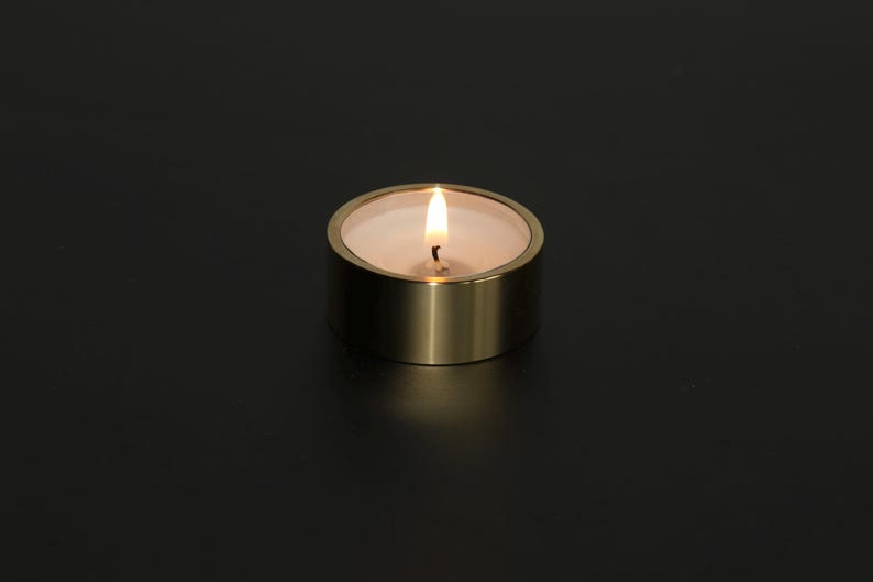 Op de afbeelding: Een brandende waxinelichtkaars in een goudkleurige metalen houder. De kaars heeft een witte was binnenkant en een kleine, flikkerende vlam. De houder is een korte cilinder met een gladde, metalen afwerking.