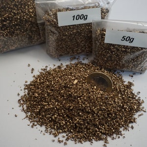 May include: Three bags of brass shavings, one labeled 100g and one labeled 50g, with a pile of brass shavings spilled out on a white surface. A coin is sitting in the pile of shavings.