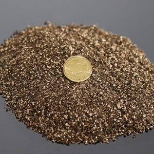 Puede incluir: Un montón de pequeñas virutas de metal de color bronce con una moneda de oro encima. La moneda tiene un diseño. Las virutas están esparcidas sobre una superficie oscura y reflectante.