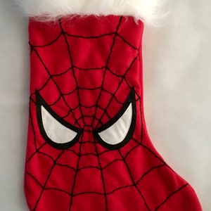 Puede incluir: Calcetín navideño rojo con diseño de Spider-Man. El calcetín tiene un fondo rojo con un patrón de telaraña negra y ojos blancos delineados en negro. La parte superior está adornada con piel sintética blanca.