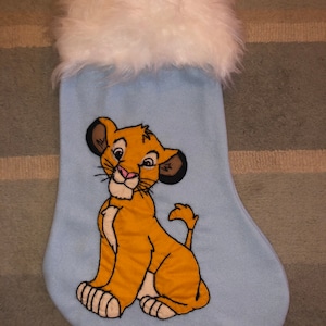 May include: A light blue Christmas stocking featuring an embroidered image of Simba from The Lion King. The stocking has a white faux fur trim at the top. The Simba image is in shades of orange, tan, and black.