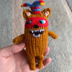 May include: A handmade, knitted toy in shades of orange, brown, red, blue, and yellow. The toy has a red mask, a blue and red hat, and a wide, toothy grin. The arms and legs are small and stubby. The toy is held in a hand.