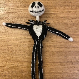 Op de afbeelding: Een handgemaakte, gebreide Jack Skellington pop. De pop heeft een witte kop met zwarte gezichtskenmerken, een zwart-wit gestreept pak en uitgestrekte armen. De pop is ongeveer 30 cm hoog en staat tegen een houten achtergrond.