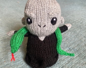 Könnte beinhalten: Handgestrickte Puppe mit grauem Kopf, großen schwarzen Augen und kleinen Ohren. Die Puppe trägt eine schwarze Robe und hat eine grüne Schlange um den Hals gewickelt. Die Schlange hat eine rote Zunge. Die Puppe ist aus Garn gefertigt.