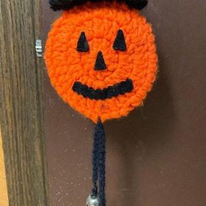Puede incluir: Una calabaza naranja de crochet con un sombrero de copa negro y una cuerda negra con cascabeles plateados que cuelgan de la parte inferior.