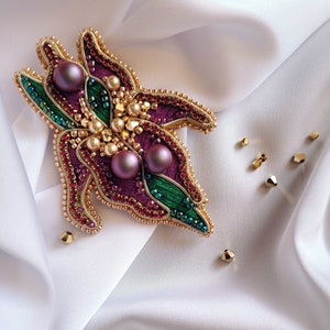 Puede incluir: Un broche bordado de color morado y verde con cuentas doradas y perlas. El broche está sobre un fondo de tela blanca.
