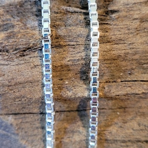Op de afbeelding: Twee zilveren kettingen met een box chain patroon, liggend op een houten oppervlak.
