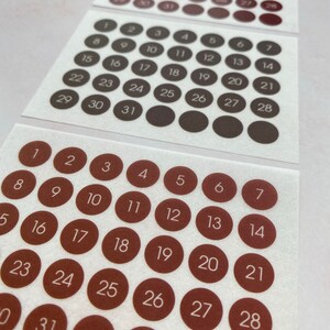 May include: Three sheets of circular stickers with numbers 1-31 printed on them. The stickers are brown with white numbers. The stickers are arranged in rows and columns, with each sticker representing a day of the month.