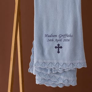 May include: A light blue knitted baby blanket with a scalloped edge, displayed on a wooden rack. The blanket features the embroidered text "Hudson Griffiths 24th April 2026" and a cross symbol.