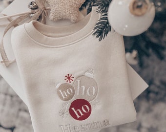 May include: A cream-colored sweatshirt with embroidered Christmas ornaments and the text "ho ho ho" in red and cream. The text "Heszna" is embroidered below the ornaments.