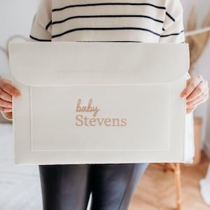 May include: Cream-colored baby keepsake box with the name "baby Stevens" embroidered in a soft, rose-gold thread. The box has a flap closure and a rectangular shape. The background is blurred, suggesting a nursery setting.