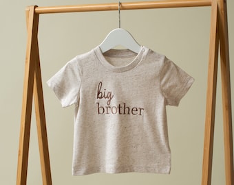 May include: A beige short-sleeved t-shirt with the words "big brother" embroidered in brown on the front. The shirt is hanging on a wooden clothes rack.