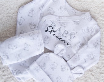 May include: A white baby outfit with a gray bear print. The outfit includes a long-sleeved onesie, pants, and a hat. The onesie has the text "BABY Stevens" embroidered on the front.