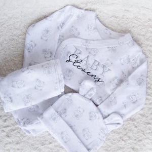 May include: A white baby outfit with a grey bear print. The outfit includes a long-sleeved onesie, trousers, and a hat. The onesie has the text "BABY Stevens" embroidered on the front.