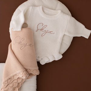 May include: A white baby sweater and a peach blanket, both embroidered with the name "Skye". The sweater has a frilled hem. The blanket has a scalloped edge. The items are on a white cushion, with a brown background.